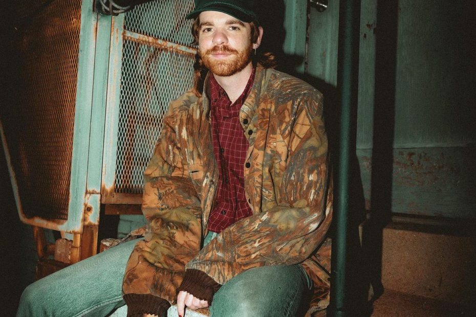 An image of the artist Field Medic sitting. He's wearing a camo jacket with a red button up shirt and a pair of jeans. In his right hand is a cigarette.