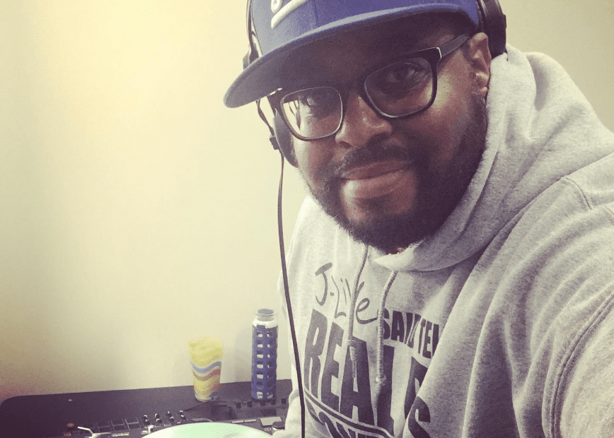 A picture of Jean Jacques Cadet, better known as J-Live. He is wearing a purple hat that says "J-Live" on it and a pair of headphones, as well as a sweater with his logo on it. He's working at his music production station and smiling at the camera.