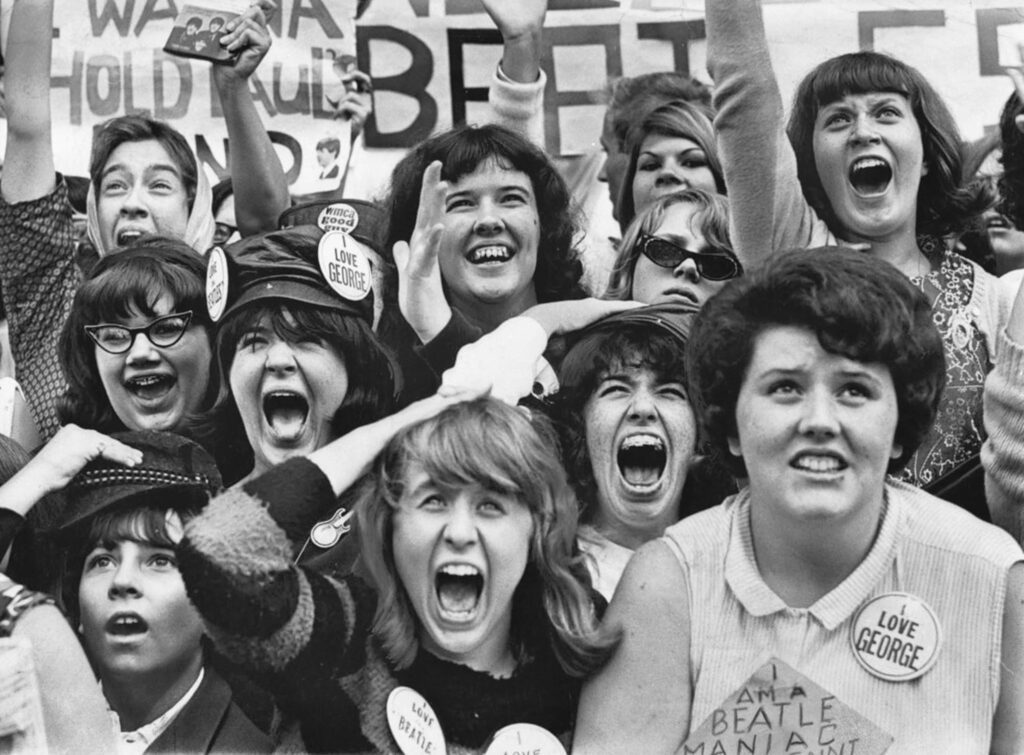 An image depicting many fans of The Beatles collected together . Many people are seen with pins saying "I Love George" and other slogans. A depiction of "Beatlemania"