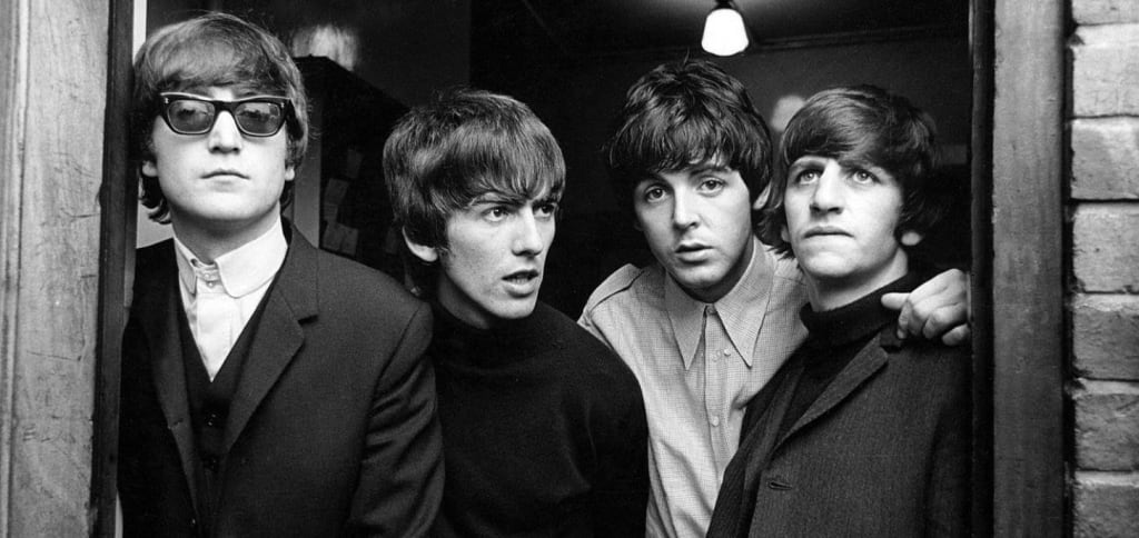 Image of the four members of The Beatles in a doorway. Paul is the only one looking at the camera.